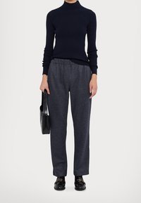 Maglione a collo alto blu navy, trama a costine; pantaloni grigi con motivo e vita elasticizzata; borsa nera in pelle; mocassini neri con accentuazione dorata.