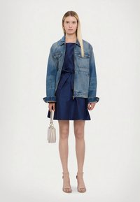 Nainen, jolla on yllään tummansininen mekko, jossa on sidottu vyötärö, suuri sininen farkkutakki, vaaleanbeige korolliset sandaalit ja joka pitää kädessään pientä valkoista käsilaukkua.