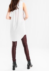 Túnica blanca sin mangas, con dobladillo asimétrico, combinada con leggings de imitación cuero burdeos y botas negras hasta el tobillo con tacón grueso.