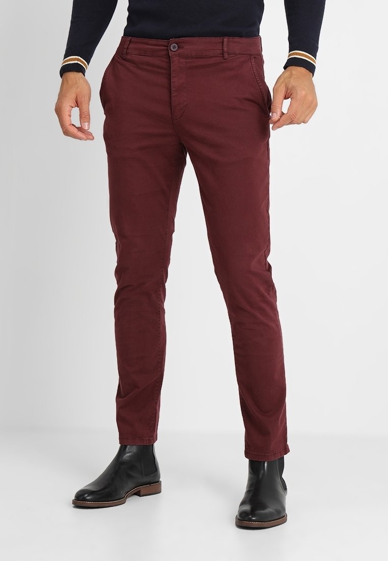 Homme portant un pantalon bordeaux slim associé à des bottines en cuir noir et un haut noir à manches longues avec des poignets rayés.