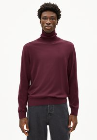 Bordeauxfarbener Rollkragenpullover aus weichem Strickstoff, mit langen Ärmeln undtaillierter Säume. Kombiniert mit dunkelgrauen Jeans.