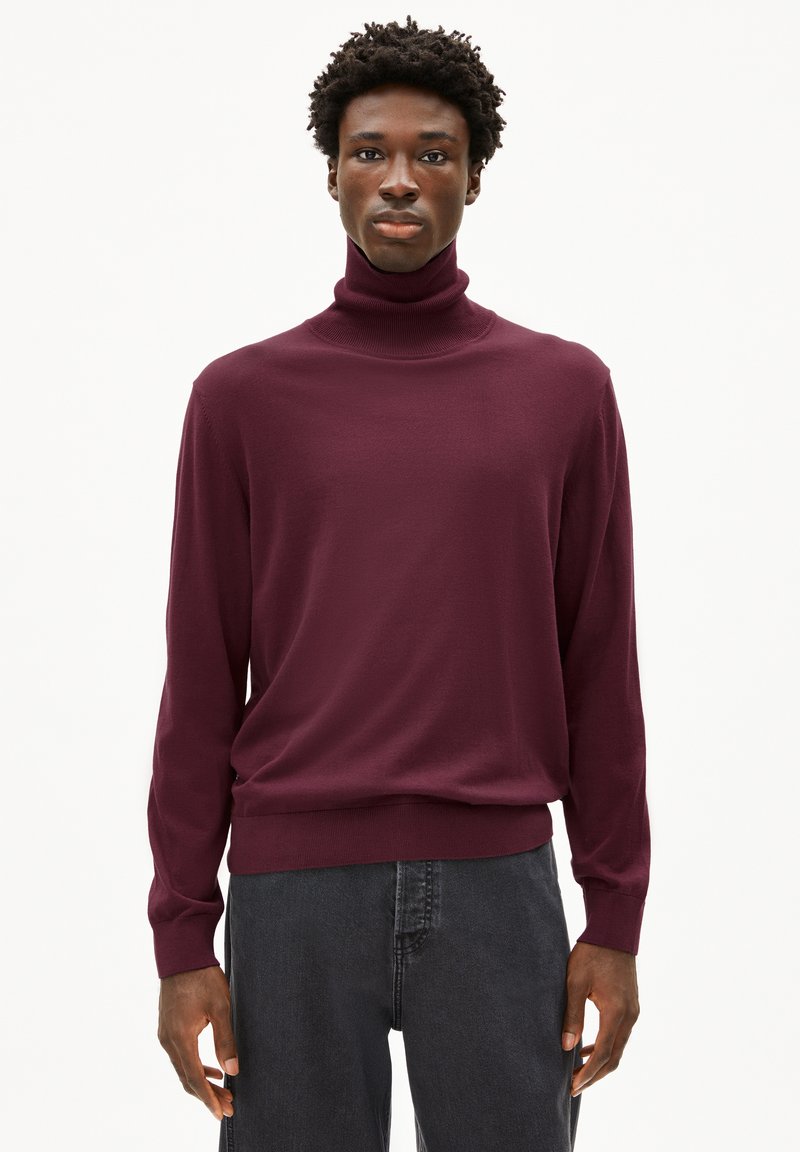 Bordeauxfarbener Rollkragenpullover aus weichem Strickstoff, mit langen Ärmeln undtaillierter Säume. Kombiniert mit dunkelgrauen Jeans.