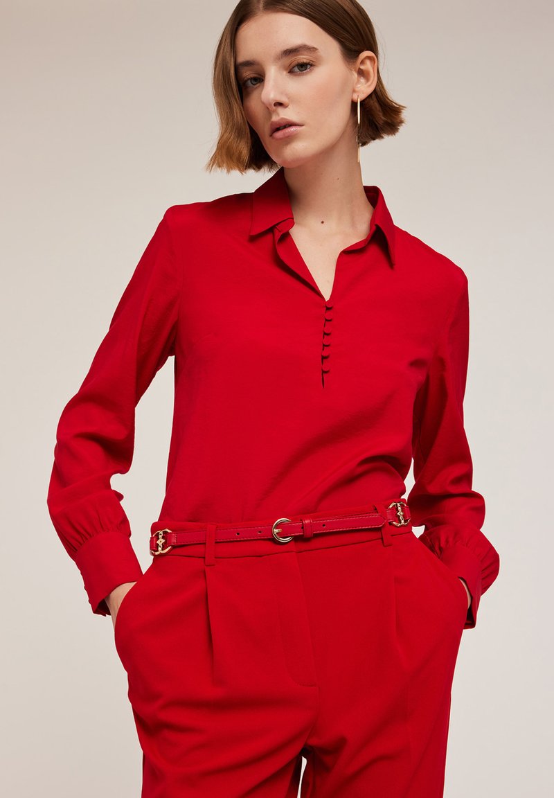 Rode blouse met lange mouwen en kraag, met een knoopsluiting en geplooide manchetten, gecombineerd met rode high-waisted broek en een bijpassende riem.