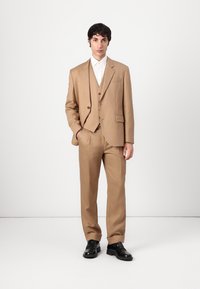 Beige three-piece suit with structured blazer, notch lapels, and matching trousers. Paired with a white button-up shirt and black shoes.