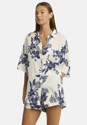 Eine Frau präsentiert ein lässiges, kurzärmliges Hemd mit Knopfleiste und floralem Navy-Muster vor einem hellen Hintergrund und strahlt eine entspannte Sommerstimmung aus.