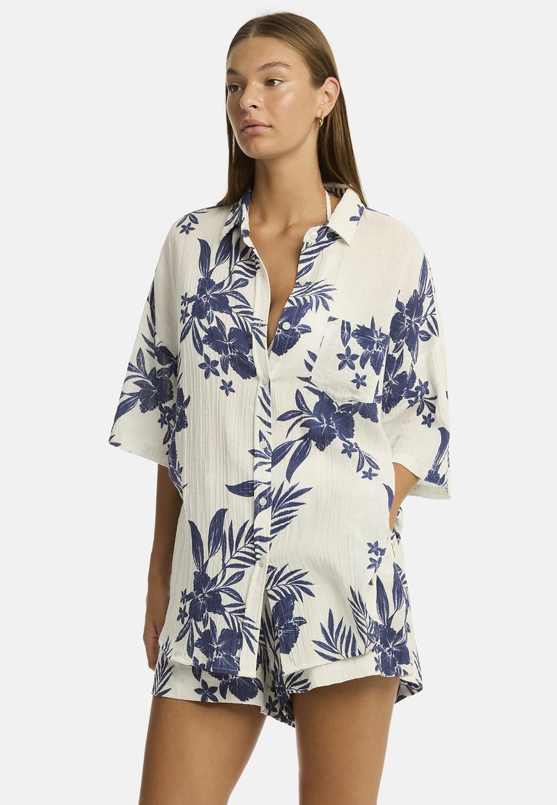 Una donna indossa una camicia button-up a maniche corte e rilassata con un motivo floreale blu su uno sfondo chiaro, trasmettendo un'atmosfera estiva informale.