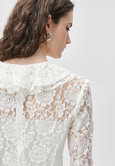 Γυναίκα με καστανά σγουρά μαλλιά, που φορά ασημένια κρίκους σκουλαρίκια και λευκό πουκάμισο από δαντέλα με λουλούδια και βολάν στο γιακά, κοιτάζοντας πλάγια.