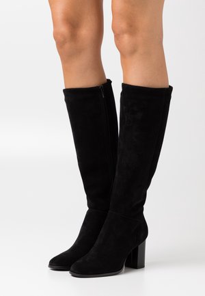 Bottes en suede noires jusqu'au genou avec des talons carrés et des zips sur les côtés, portées sur des jambes nues sur un fond blanc uni.