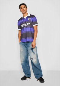 Lilla og grå stribet fodboldtrøje med hvid krave og "REPLAY"-logo, parret med slidte blå bredbenede jeans og sorte sneakers.