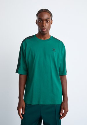 Grön bomulls t-shirt med avslappnad passform, utrustad med svarta randiga detaljer på axlarna och en liten svart logotyp på bröstet.
