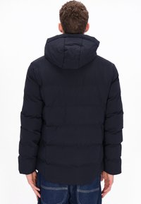 Sötétkék puffertalpú dzseki kapucnival, melyet steppelt mintázat és elasztikus mandzsetták jellemeznek. Az anyag simának és szigeteltnek tűnik.