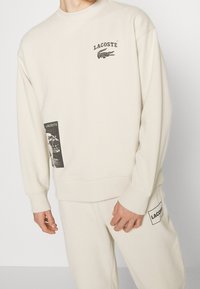 Ensemble sweat-shirt et pantalon beige Lacoste avec logos noirs et un imprimé graphique sur le bas gauche du sweat-shirt.