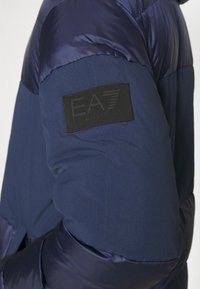 Granatowa kurtka puchowa z kontrastującymi błyszczącymi i matowymi fakturami. Czarna gumowana naszywka z logo na rękawie. Smukła sylwetka.