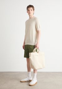 Maglione beige a maniche corte in maglia, pantaloni corti verdi, sneakers bianche con suole marroni e una grande borsa tote in tela beige. Texture liscia.