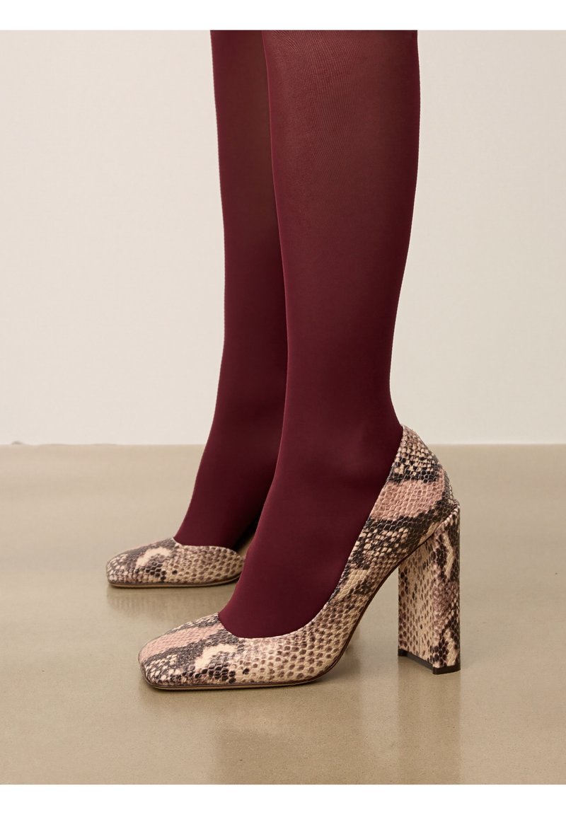 Escarpins à talons hauts avec un motif de serpent rose et brun, présentant un bout carré et un talon épais. Portés avec des collants bordeaux.