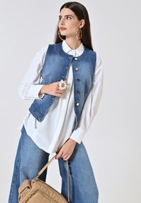 Gilet di jeans con colletto e chiusura a bottoni, indossato sopra una camicia bianca. Presenta grandi orecchini e una borsa beige con motivo a trapunta.
