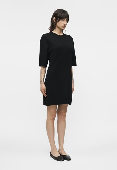 Γυναίκα με σκούρα μαλλιά που φορεί απλό μαύρο φόρεμα μέχρι το γόνατο με μανίκια μέχρι τους αγκώνες και μαύρες φλατ παπούτσια, στέκεται μπροστά σε λευκό φόντο.