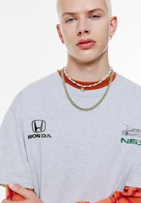 Värillä vaalea harmaa t-paita, jossa on musta "HONDA"-logo, oranssi kaulus ja vihreä "NSX"-grafiikka. Yhdistettynä kerroksellisiin hopea- ja helmikaulaketjuihin.