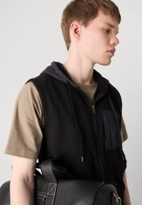 Μαύρος αμάνικος φορώντας μια γκρίζα κουκούλα, μπροστινές τσέπες και φερμουάρ, φορεμένος πάνω από μια μπεζ μπλούζα; κρατώντας μια μαύρη τσάντα με υφή.