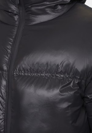 Zbliżenie na czarną pikowaną kurtkę o błyszczącej powierzchni, z kapturem i przednim zamkiem, z wytłoczonym logo "GOOD FOR NOTHING" na piersi.