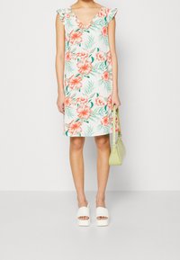 Blumenkleid mit weißem Hintergrund, das rosa Hibiskusblüten und grüne Blätter zeigt. Rüschenkragen und gerader Schnitt. Getragen mit weißen Blockabsatzsandalen und einer hellgrünen Handtasche.