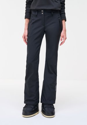 Personne portant un pantalon de neige noir avec des poches zippées et des bottes d'hiver noires, debout devant un fond uni.