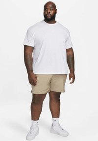 Man med tatueringar iklädd en vit Nike t-shirt, beiga shorts, vita strumpor och vita sneakers, stående mot en ren vit bakgrund.