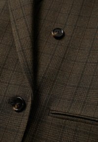 Ruitblazer van bruin stof met een gestructureerde weving, voorzien van twee zwarte knopen en een enkele borstzak. Plat uitzicht, met nadruk op de details.