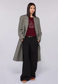 Šedý vlněný kabát přes bordó svetr s logem, kombinovaný s černými širokými kalhotami a silnou podešví šitými botami v bordó a béžové barvě.