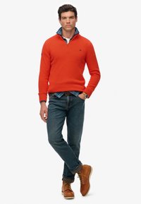 Maglione arancione a maglia con colletto a zip, indossato sopra una camicia a quadri blu. Abbinato a jeans blu e scarponcini marroni con lacci.