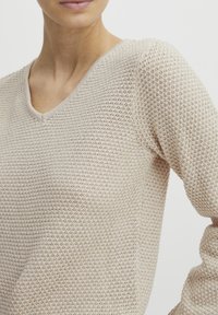 Pull beige avec un motif en tricot texturé, doté d'un col en V et de manches longues. Le tissu semble léger et respirant.
