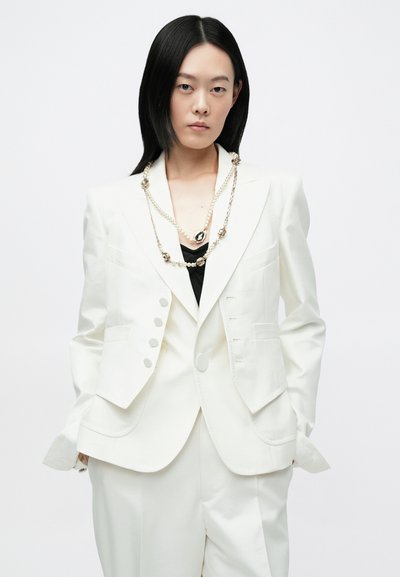 Witte getailleerde blazer met ingezette revers, vier knopen aan de voorkant en klepzakken, gecombineerd met een zwarte top en een gelaagde ketting.