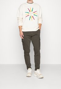 Off-weißes Sweatshirt mit mehrfarbigem Sonnen-Gesichts-Motiv, kombiniert mit olivgrünen Cargo-Hosen. Weiß-beige Sneakers runden den Look ab.