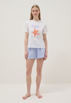 Frau steht barfuß, trägt ein weißes T-Shirt mit orangem Seestern und blauem Text, kombiniert mit hellblauen Shorts vor einfarbigem Hintergrund.