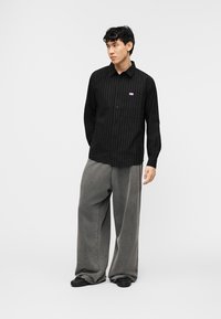 Νεαρός άνδρας που στέκεται φορώντας μαύρο πουκάμισο με λεπτές ρίγες και κουμπιά, φαρδιά γκρίζα παντελόνια και μαύρα παπούτσια, σε απλό φωτεινό φόντο.