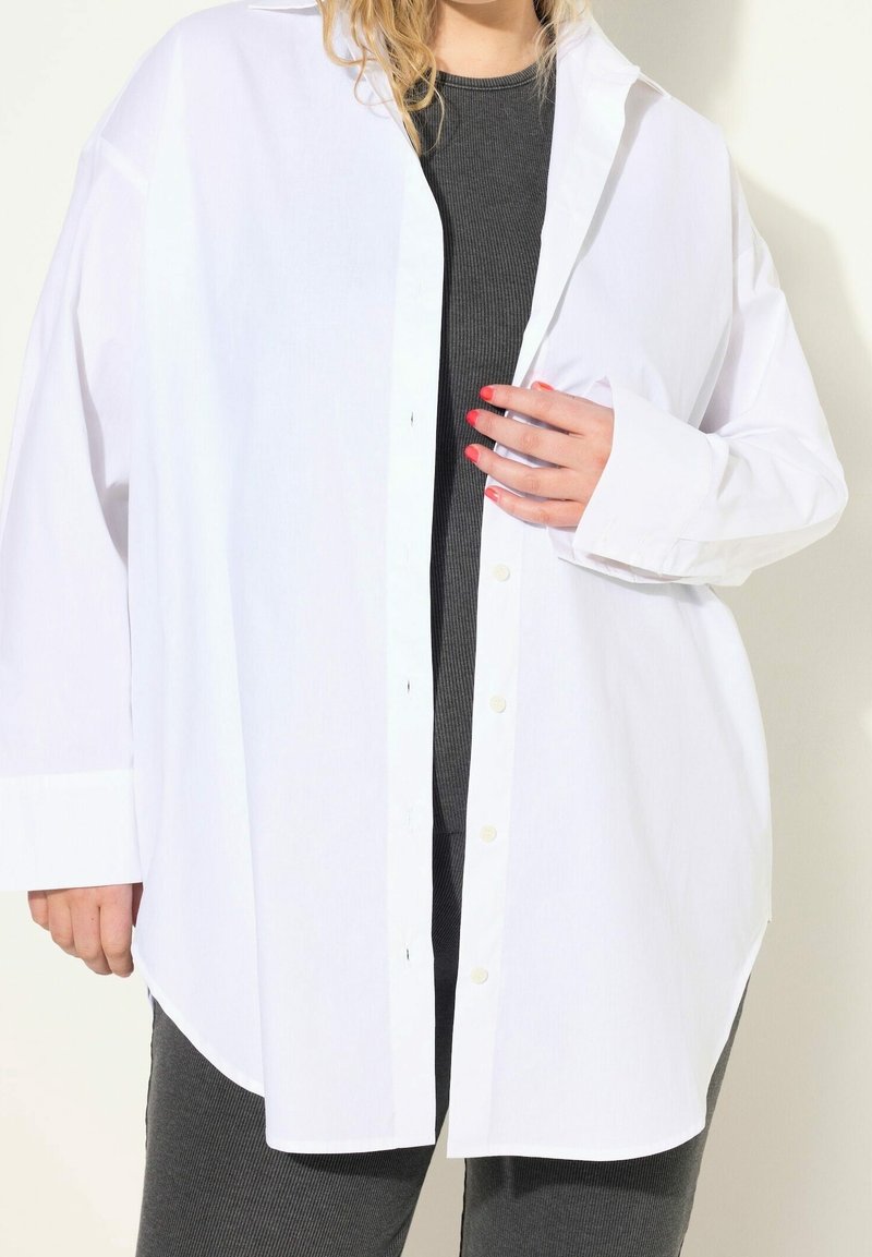 Personne portant une chemise blanche oversize boutonnée sur un haut côtelé foncé et un pantalon, une main touchant le devant de la chemise montrant un vernis à ongles rouge.