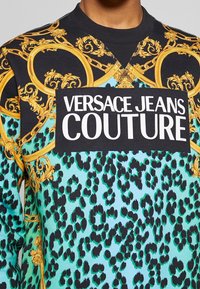 Sudadera gráfica con estampado de leopardo turquesa y patrón barroco dorado. El logotipo central dice "VERSACE JEANS COUTURE" en blanco y negrita.
