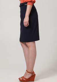 Jupe marine longueur genou avec une texture lisse, dotée d'une ceinture à la taille. Associée à des sandales compensées rouge orangé.