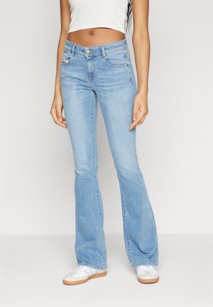 Femme portant un jean bootcut bleu clair, un crop top blanc et des baskets blanches, debout devant un fond uni.