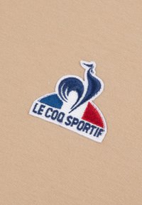 Vyšiované logo na béžové tkanině. Design zahrnuje stylizovaného kohouta s modrými, bílými a červenými akcenty, spolu s textem "LE COQ SPORTIF."