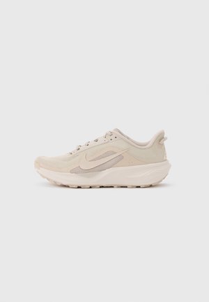 Zapatilla de running ligera beige con detalles de malla, suela acolchada y logo de Nike swoosh en el lateral, mostrada de perfil sobre un fondo liso.