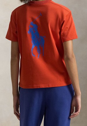Persoon die een oranje T-shirt draagt met een grote blauwe polo-speler afbeelding op de achterkant en een blauwe broek tegen een neutrale achtergrond.