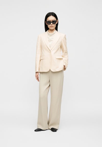 Femme portant un blazer crème, pantalon large assorti, lunettes de soleil noires, chaussures noires, cheveux noirs lisses sur un fond uni.