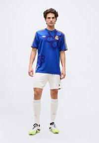 Young man standing in blue floral-patterned sports jersey, white shorts, knee-high socks, and cleats against a plain white background.