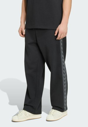 Person trägt schwarze Jogginghose mit seitlichen, mit Logo versehenen Streifen und weiße Sneakers, steht vor einem einfarbigen hellen Hintergrund.