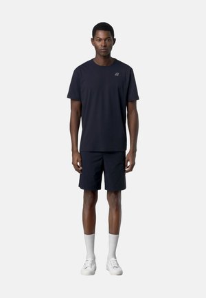 Uomo in piedi frontale che indossa una maglietta nera, pantaloncini neri, calzini bianchi e scarpe da ginnastica bianche su uno sfondo chiaro uniforme.