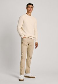 Kremasti rebrasti pulover, bež uske hlače i bijele tenisice s smešnim potplatima, stojeći pred svijetlom sivom pozadinom.