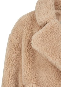Beige faux shearling ülikond, millel on pehme, tekstuuriga pind, laiad sagsid ja lõdvem istuvus, ilma nähtavate kinnitusvahendite või kaunistusteta.