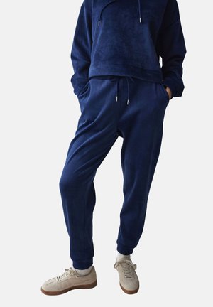 Person iført navyblå ribstrikkede joggebukser og matchende hættetrøje, kombineret med beige sneakers og hvide sokker, stående med hænderne i lommerne.