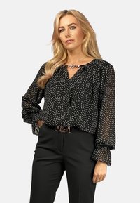 Schwarze Bluse mit weißen herzförmigen Mustern, fließenden Ärmeln und einem Kettendetail am Ausschnitt. Kombiniert mit schwarzen Hosen.